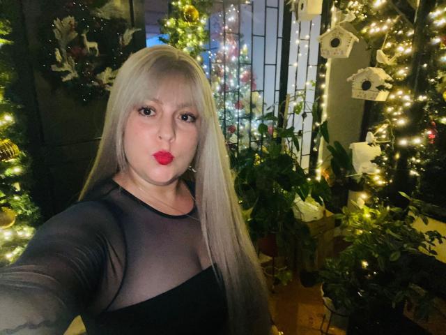 SamanthaGold with long blonde hair, red lipstick, and a sheer black top, showing off her boobs in a festive, Christmas-lit room with decorated trees and plants.