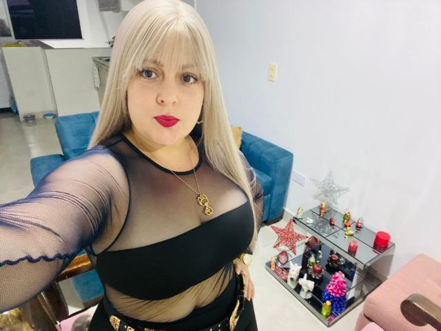 SamanthaGold, con lunghi capelli biondi, rossetto rosso, una maglia nera trasparente che mette in mostra il suo seno prosperoso, un paio di pantaloni neri a fascia, una collana d'oro, in piedi in una stanza con una sedia blu e una mensola..