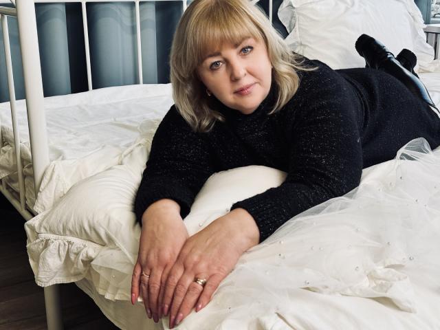 MilenaWendy with blonde hair, black sweater, lying on white bed, hands in front, ring on finger, black boots, white pillows, metal bed frame, smiling, fair skin, medium tits.