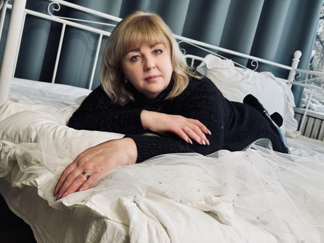 MilenaWendy, blonde, lying on white bed, black sweater, hands on pillow, ring on finger, blue eyes, slightly smiling, white metal bed frame, white pillows, sheer white bedsheet.