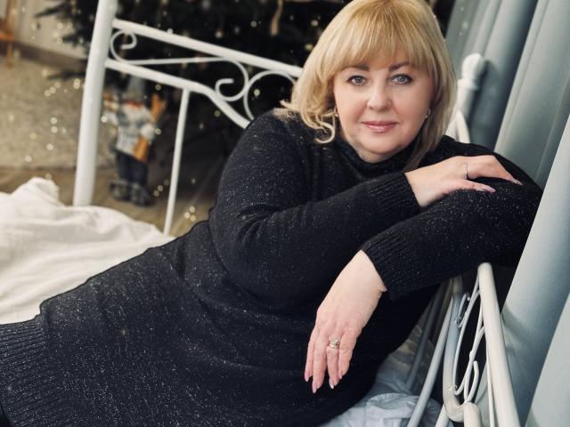 MilenaWendy with blonde hair, blue eyes, black sparkly sweater, lying on white bed, right arm on bed, left arm on window, ring on finger, subtle smile.