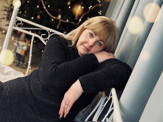 MilenaWendy with blonde hair, wearing a black glittery sweater, leans on a white metal bed, eyes blue, smiling softly, hands resting, ring visible, Christmas tree lights in background.