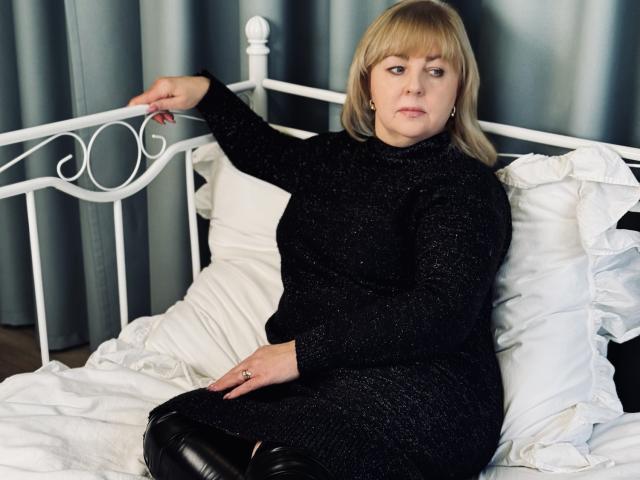 MilenaWendy with blonde hair, black sparkly dress, and black leather pants, sitting on a white bed, arm raised, looking annoyed, white metal bed frame, white pillows, gray curtains.