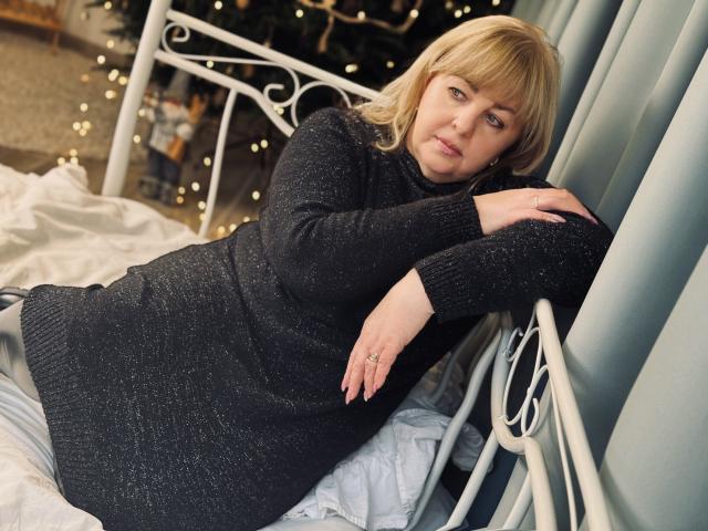 MilenaWendy with blonde hair, wearing a black sparkly sweater, lying on a white bed, arms resting on a metal headboard, looking at the camera, pale skin, ring on right hand.