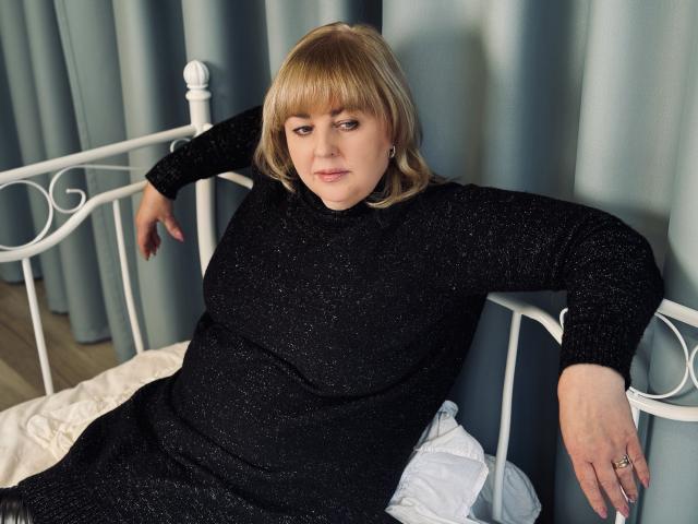 MilenaWendy, chubby with blonde bob, wearing black sparkly dress, sitting on white bed, arms resting, looking slightly annoyed, blue curtains in background, wooden floor, ring on right hand.