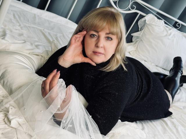 MilenaWendy is lying on a bed, wearing a black sparkly top, black boots, and white sheer fabric, with her hand on her cheek, looking at the camera.