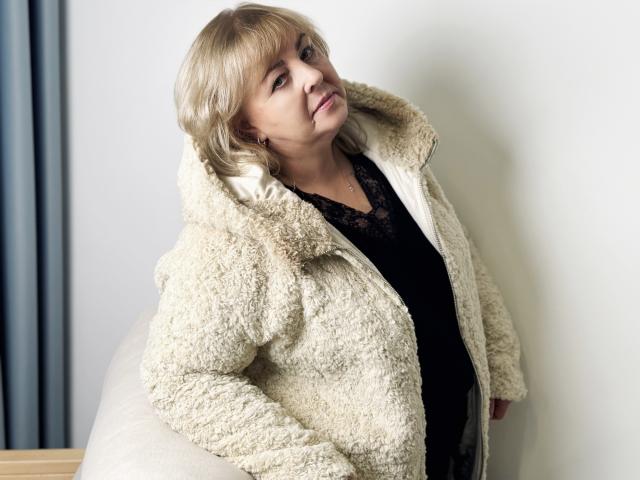 MilenaWendy with blonde bob, wearing a fluffy white coat, black lace top, leaning against a white wall, smiling slightly, looking relaxed, coat's texture is soft and fuzzy.