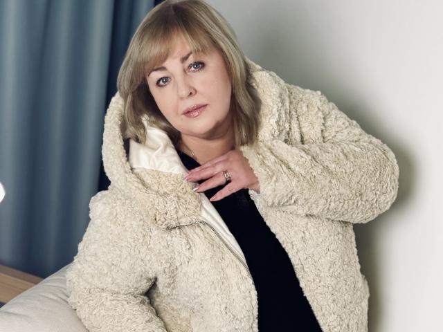 MilenaWendy with blonde hair, blue eyes, wearing a fluffy white coat, black top, and a ring, leaning against a white wall, looking at the camera, slightly smiling.