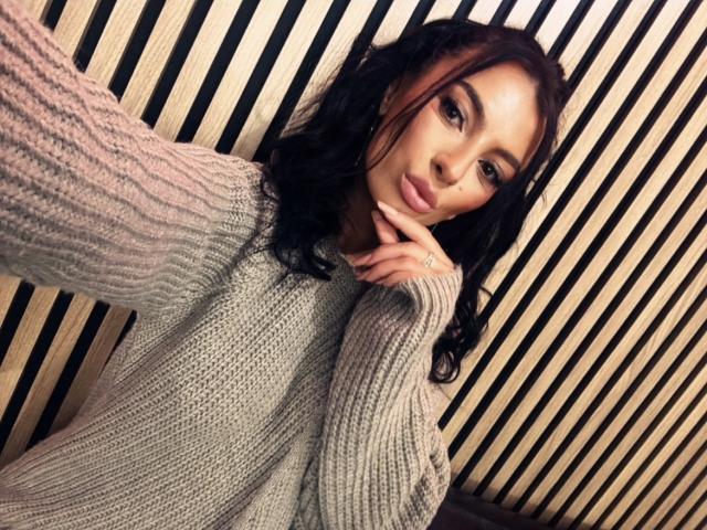 KinkyAmelie with dark wavy hair, wearing a gray knit sweater, has a ring on her finger, and a sexy, pouty expression, looking into the camera with a slight smile.