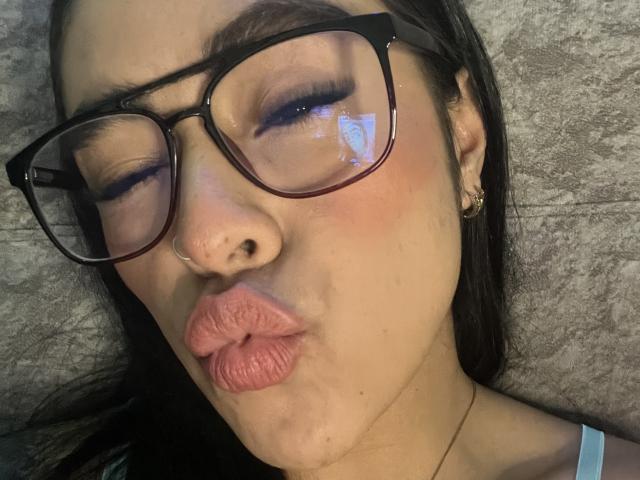 StellaBeckker with pouty pink lips, black-framed glasses, and a nose ring, eyes closed, reflection in glasses, wearing a tiny blue top, lying on a grey textured surface.