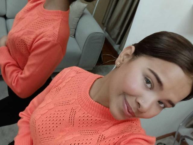 ChloeVixen with medium tits in an orange knit sweater, smiling slightly, brown hair in a ponytail, green eyes, nose piercing, small earring, standing in a room.