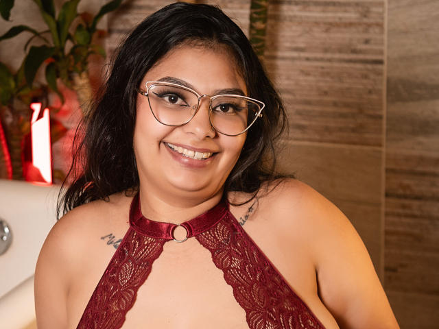 JulietaOwen with dark hair, glasses, smiling, wearing a red lace halter top, showing small tattoos on her shoulders, her tits visible, in a bathroom.