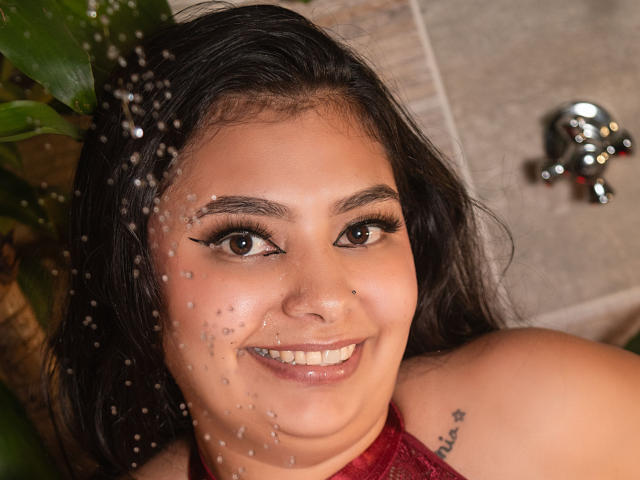 JulietaOwen's face with water droplets on her skin, smiling, brown eyes, dark hair, red halter top, small nose piercing, tattoo on her right shoulder, in a shower.