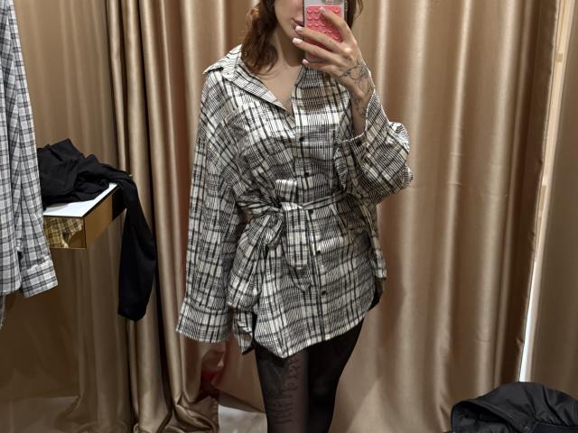 SensualMila in a plaid shirt dress, black tights, holding a pink phone, mirror selfie, beige curtain background, black shirt on a shelf to the left.
