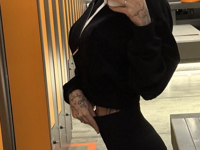 SensualMila's side profile in a black hoodie and tight black pants, showing tattoos on her hands, with her phone in the other. Her tits are covered by the hoodie.