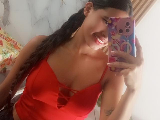 ScarlettRoss69 with long black hair, red cleavage top, smiling, holding pink phone with colorful case, showing small tattoo on left arm, medium tits, light brown skin.