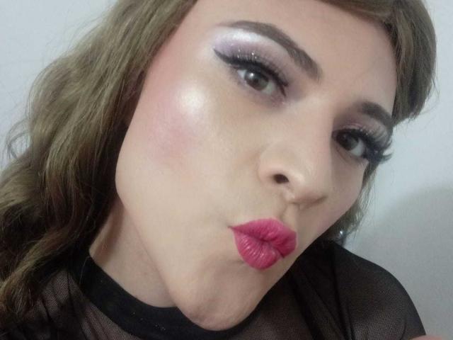 CattaSmithTs avec rouge à lèvres rose, lèvres pulpeuses et fard à paupières violet brillant, portant un haut en maille noire, des cheveux bruns ondulés et un eye-liner noir épais, regardant la caméra avec une expression sexy..
