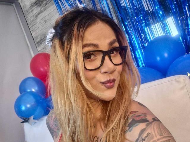 AzhunaStar with blonde hair, black glasses, and tattoos, looking at the camera with a pouty expression, surrounded by blue and red balloons.