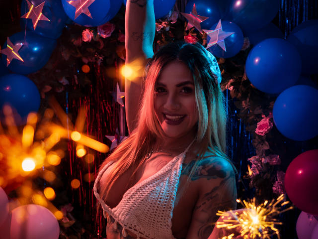 AzhunaStar with blonde hair, smiling, arm raised, wearing a white crochet top, exposing her tits, with tattoos on her arm, surrounded by blue balloons and star-shaped decorations.