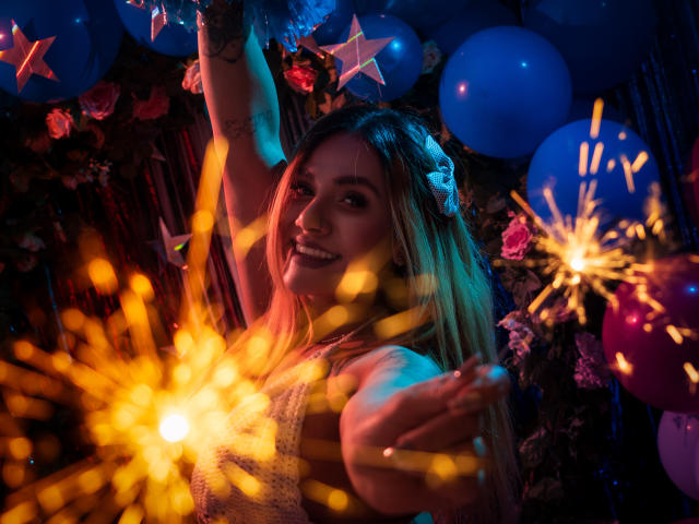 AzhunaStar with blonde hair, wearing a white dress, holding sparklers, smiling, blue balloons, and star decorations in the background, under colorful, festive lighting.
