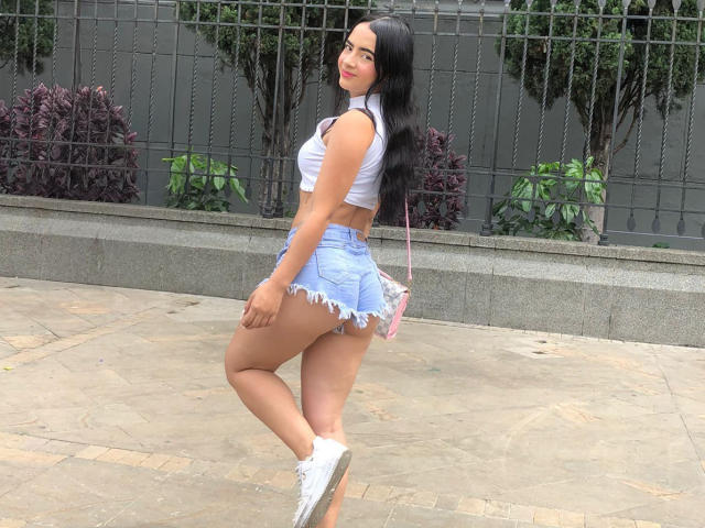KonanStar has long black hair, a white crop top, and frayed blue denim shorts showing her round ass. She's standing on a stone patio in white sneakers.