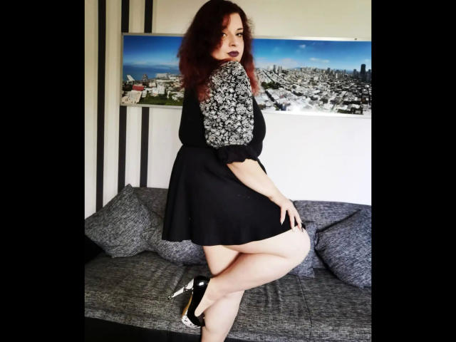 CashladyJacky97 - aantrekkelijk met donkerrood haar, een zwart-wit bloemen top, een zwarte rok en zwarte hoge hakken, waarbij haar blote billen zichtbaar zijn. Ze staat op een grijze bank en kijkt om..