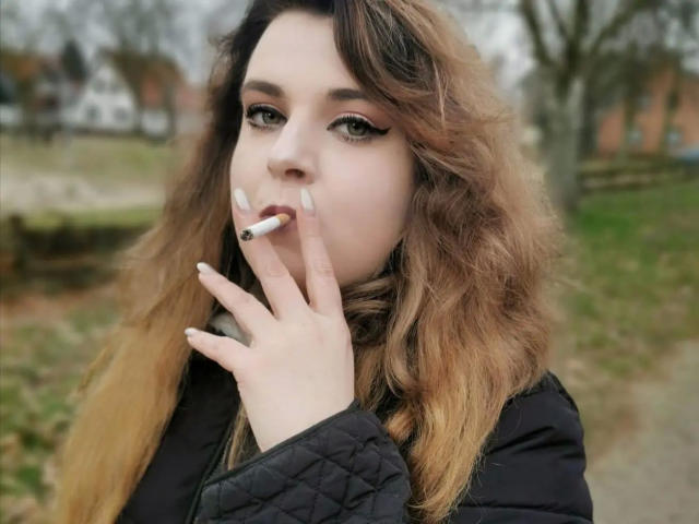 CashladyJacky97 - Une femme séduisante avec de longs cheveux bruns ondulés, portant une veste noire matelassée, fumant une cigarette, qu'elle tient près de sa bouche, avec des ongles vernis en blanc et des yeux verts..