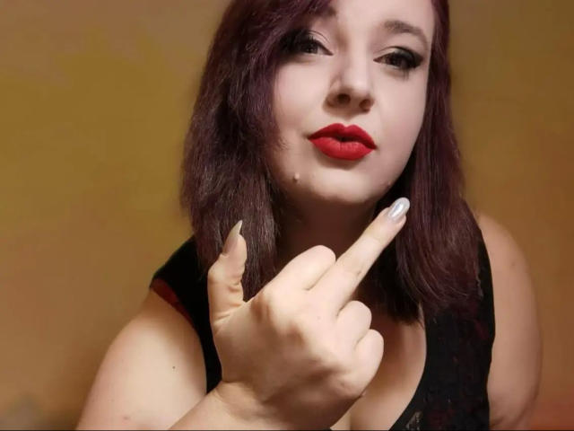 CashladyJacky97-belle, avec du rouge à lèvres, des cheveux violet foncé et une peau pâle, pointant du doigt avec son index, portant un haut noir sans manches, mettant en valeur ses ongles peints en blanc..