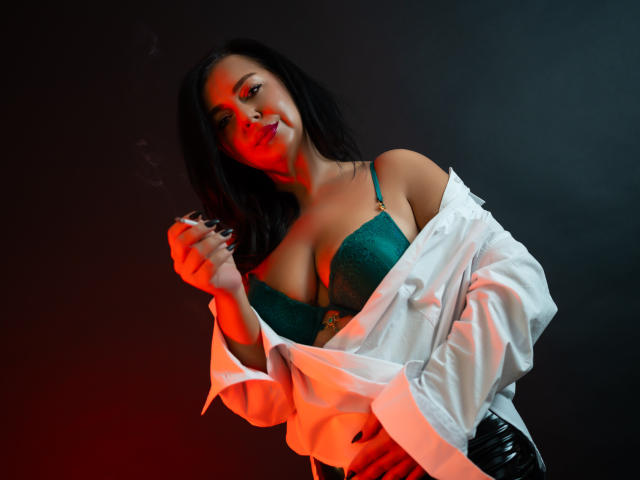 MonicaVixen with dark hair, green lace bra, and white shirt off-shoulder, holding cigarette, red and orange light, showing her tits, black background, and red lipstick.