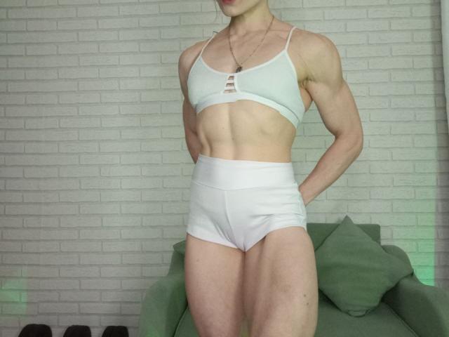 VeraKyk with muscular, defined abs wearing a white sports bra and high-waisted white underwear, hands on hips, green armchair in background, white brick wall.