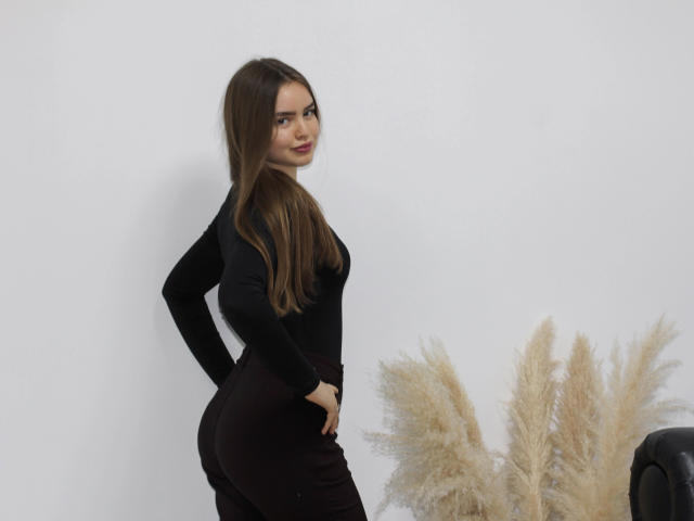 MiaCalhoun with long brown hair, black long-sleeve top, and dark pants, standing sideways, hand on hip, looking over shoulder, ass prominent, puffy beige grasses in foreground.