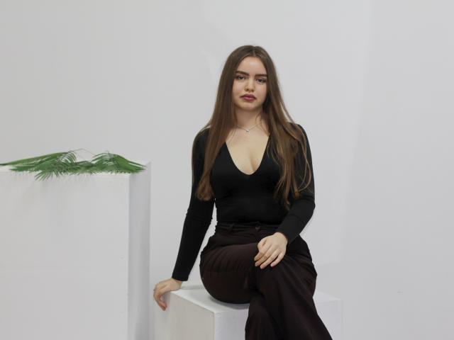 Mia Calhoun, con lunghi capelli castani, indossa una maglia nera a V, pantaloni marroni, è seduta su un cubo bianco, con le mani in grembo, a sinistra c'è una pianta verde, sullo sfondo c'è il bianco, e guarda leggermente verso la telecamera..