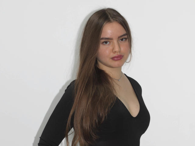 MiaCalhoun with long brown hair, wearing a black V-neck, showing small tits, subtle ass, and a necklace against a white background. She has a serious expression and red lipstick.