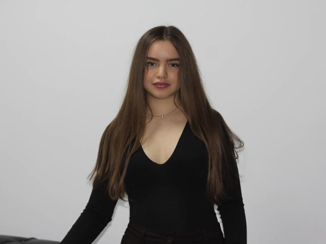 MiaCalhoun with long brown hair, wearing a black V-neck top, showing her small tits and a silver necklace, standing against a plain white wall.