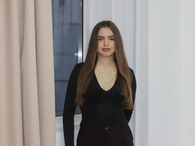 Mia Calhoun è in piedi, con lunghi capelli castani, indossa una maglia nera a maniche lunghe con un profondo scollo a V e pantaloni scuri, mostrando leggermente i suoi seni attraverso la maglia..