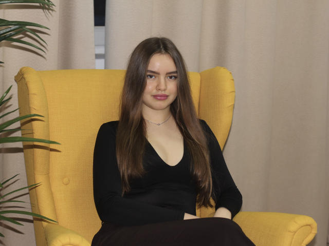 Mia Calhoun, con lunghi capelli castani, una maglia nera a V e rossetto rosa, seduta su una sedia gialla, con le braccia incrociate, un sorriso appena accennato, indossa una sottile collana e appare sicura di sé..
