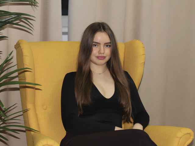 Mia Calhoun, con lunghi capelli castani, indossa una top nera a V profondo, ha un seno piccolo, è seduta su una poltrona gialla, con le braccia incrociate, un sorriso appena accennato, una gonna nera, tende beige, una pianta verde a sinistra..