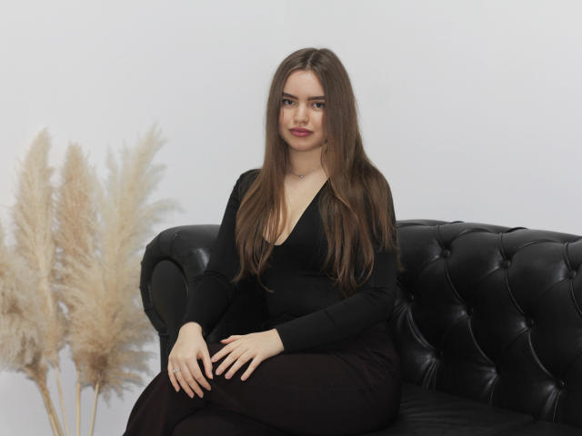 MiaCalhoun with long brown hair, black V-neck top, and black pants, sitting on a black leather couch, hands on lap, puffy beige grasses to the left.
