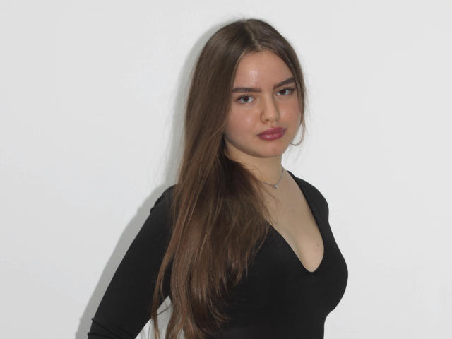 MiaCalhoun with long brown hair, black deep V-neck top, small chest, fair skin, subtle makeup, and a slight smirk, standing against a plain white background.