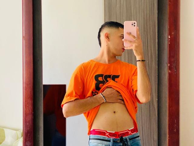 BaironSinclair in an orange shirt, lifting it to show his flat, tattooed belly with a navel piercing, wearing blue jeans and red underwear, taking a mirror selfie.