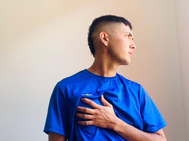 BaironSinclair with short, dark hair, blue t-shirt, and hand on chest, looking to the side, showing a slight pensive expression.