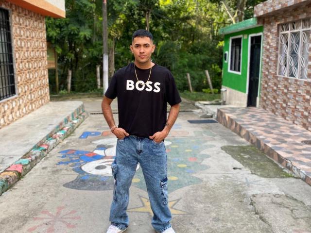 BaironSinclair stands in a concrete alley, wearing a black "BOSS" shirt, blue jeans, and white sneakers, with hands in pockets, looking at the camera.