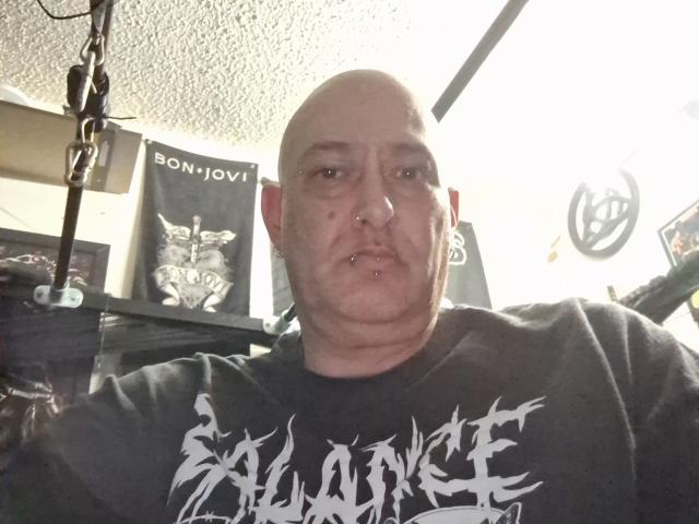 PiercedColck, bald with a nose ring and lower lip piercing, wearing a black "Slayer" shirt, has a chubby face and a neutral expression. Ceiling light visible.