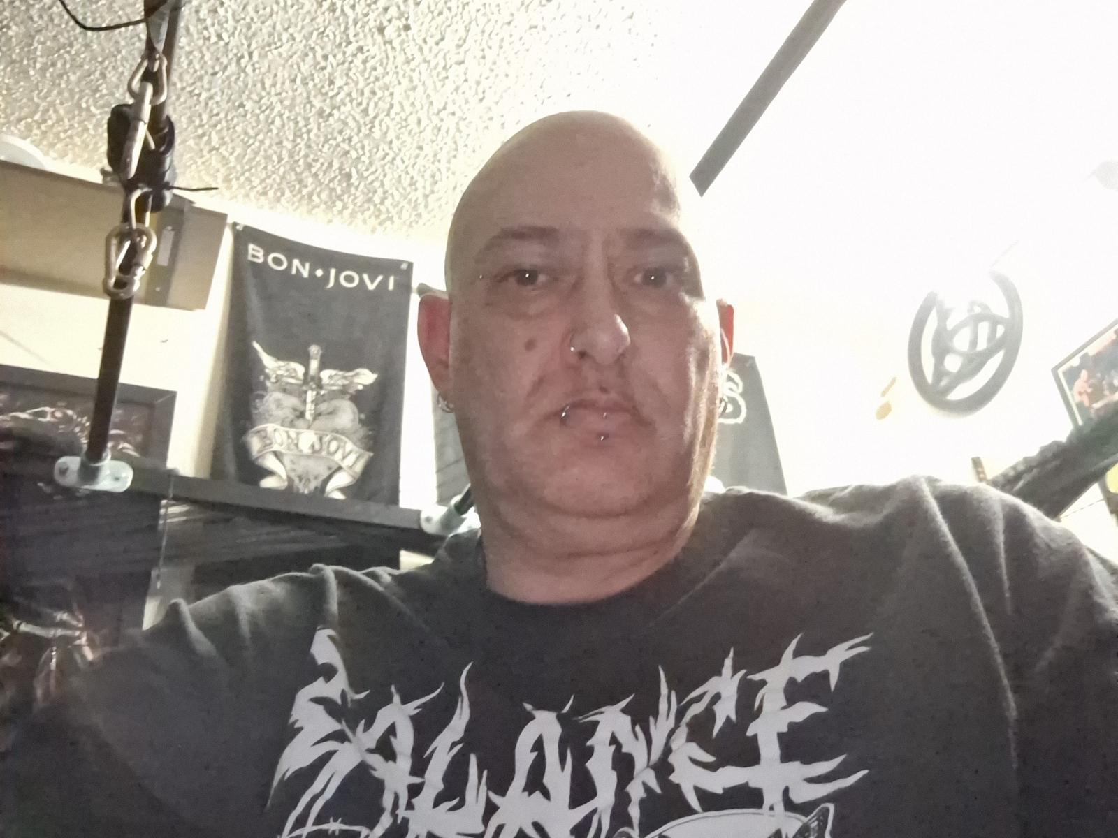 PiercedColck, bald with a nose ring and lower lip piercing, wearing a black "Slayer" shirt, has a chubby face and a neutral expression. Ceiling light visible.
