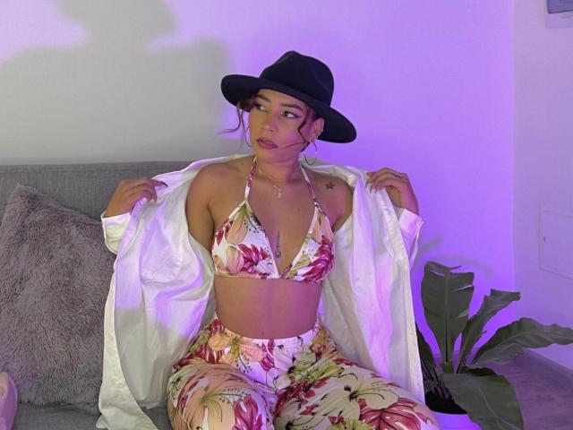 SaharaArisa with floral bikini top, matching pants, and open white shirt, wearing a black hat, sitting on a gray couch, purple lighting, and a plant in the background.