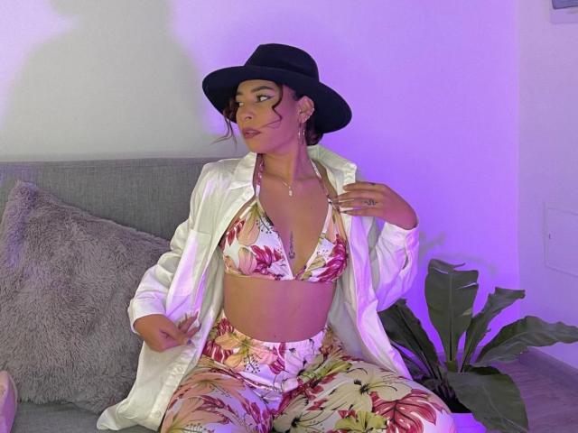 SaharaArisa with tan skin, black hat, white shirt, floral bikini top, and matching pants, sitting on a gray couch, purple light, looking to the side, hands on shirt.