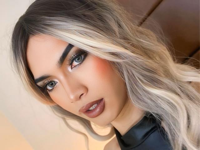 AmandaMooree's face with blonde, wavy hair, thick eyebrows, green eyes, dark makeup, and glossy brown lips. Her tits are covered by a black, leather-like top.