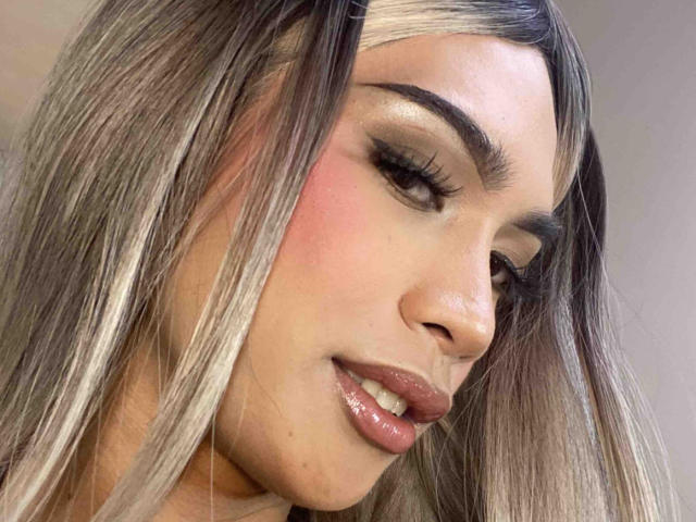 AmandaMooree's face with long, straight blonde hair, thick eyebrows, and glossy pink lips, looking down with a slight smile. Her nose is small, and her skin is fair.