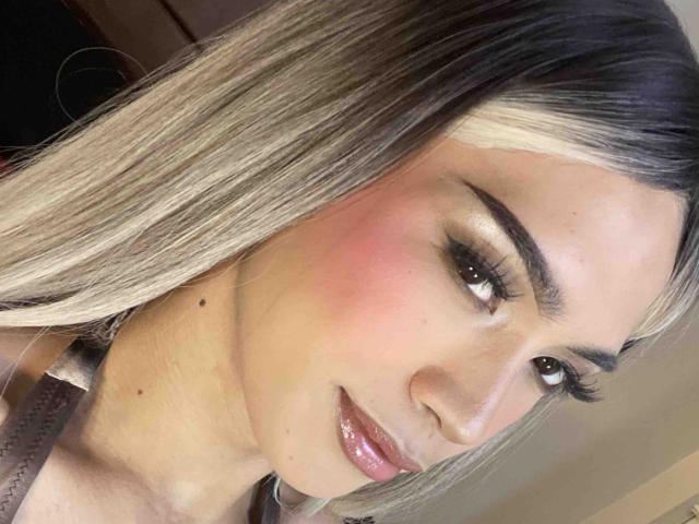 AmandaMooree's face, with blonde-to-dark-brown ombre hair, pink blush, glossy pink lips, and thick eyelashes, smiling slightly. She has small moles on her cheek.
