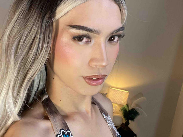 AmandaMooree with blonde hair, pink blush, glossy pink lipstick, and subtle eye makeup, wearing a grey halter top, looking at camera with slightly parted pink lips.